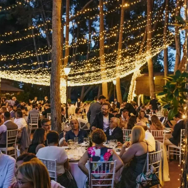 Cena al aire libre con numerosos invitados bajo un techo de luces cálidas entre los árboles.