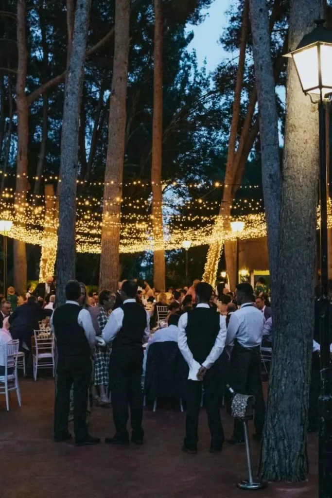 Cena al aire libre con numerosos invitados bajo un techo de luces cálidas entre los árboles